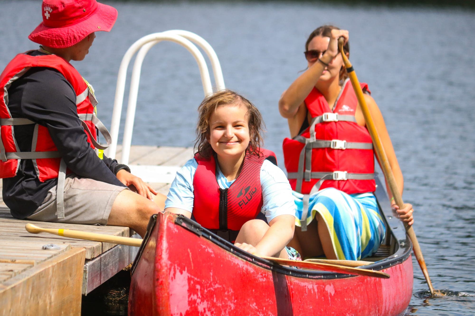 A camper smiles from a canoe at the dock while a lifeguard and a counsellor look on.